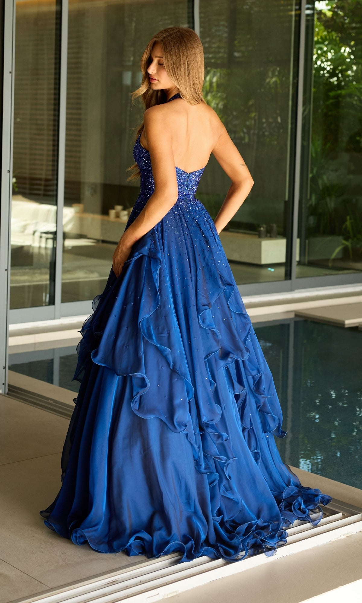 A woman stands indoors near a pool, wearing the Ava Presley 42235: a long halter A-line formal dress.
